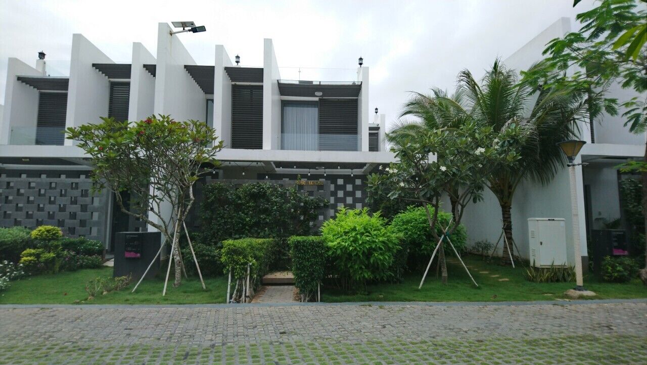 Un bâtiment moderne à la façade symétrique, doté d’une entrée pavée et d’un jardin bien entretenu, agrémenté de diverses plantes et d’un petit pont.