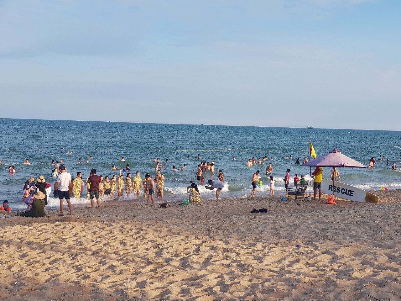 Ein überfüllter Strand mit badenden und Menschen, die am Sand stehen, einem Rettungsschwimmerstand und einem Rettungsbrett.