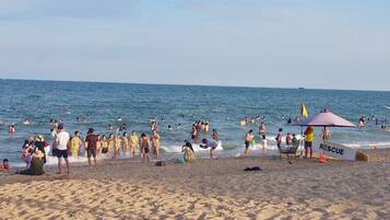 Ein überfüllter Strand mit badenden und Menschen, die am Sand stehen, einem Rettungsschwimmerstand und einem Rettungsbrett.