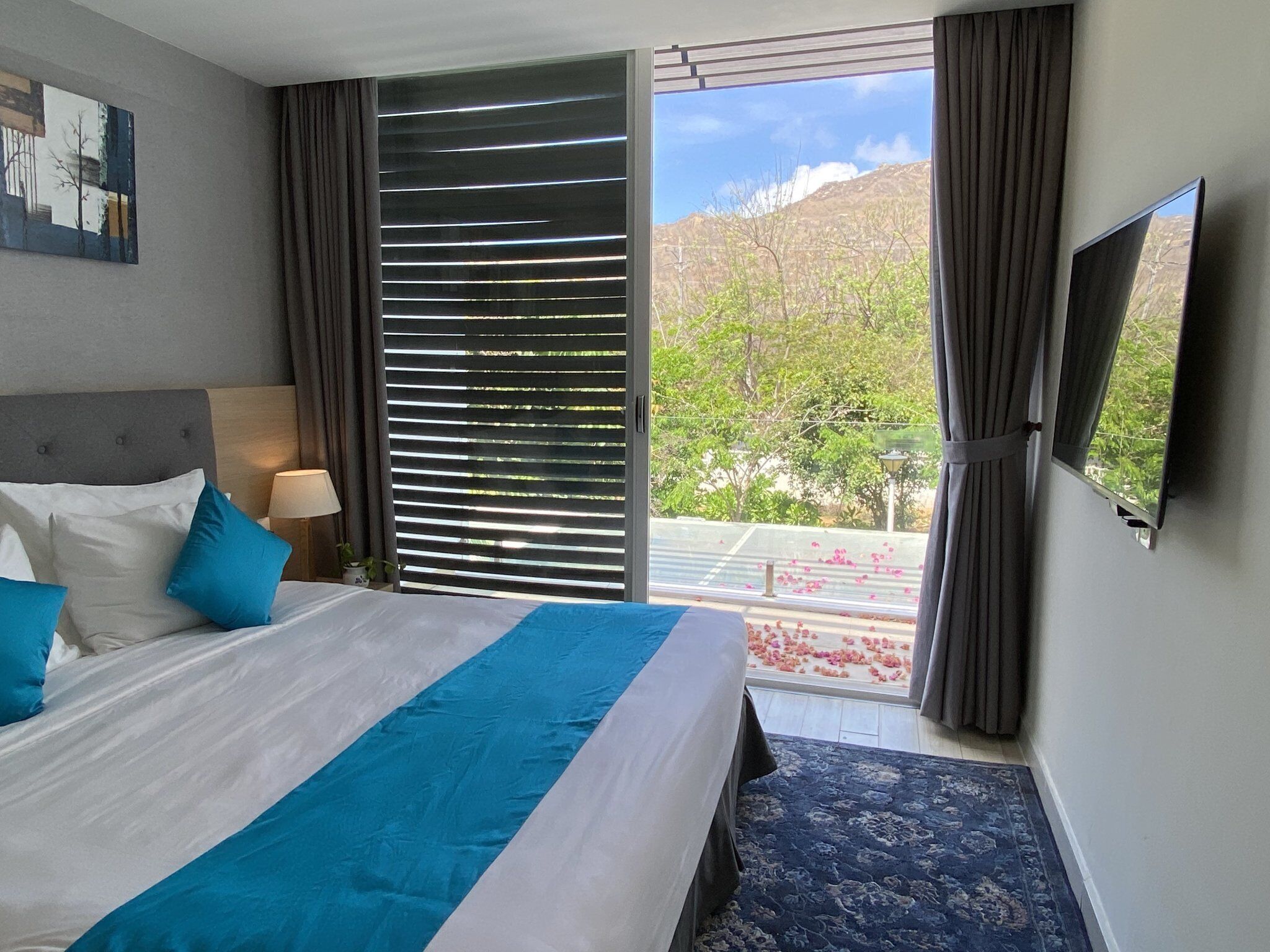 A hotel room with a large bed, a flat-screen TV, a window with blinds, and a view of greenery outside.