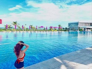 A swimming pool with clear water, surrounded by lounge chairs and umbrellas, and a modern building in the background. - Villa Bien - Resort Oceanami (Ho Chi Minh City)