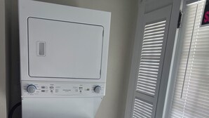 A white wall-mounted washer and dryer unit with control knobs and a small pink label, located in a corner next to a door with white blinds. - Peace Apartment, upstairs with king size bed. (Anniston)