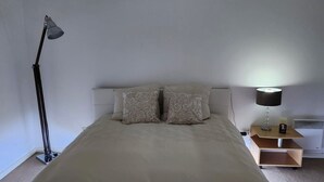 Ein Bett mit weißer Bettdecke und zwei Kissen mit Muster, eine Nachttischlampe und ein kleiner Holfnachtisch mit Lampe und Kerze.