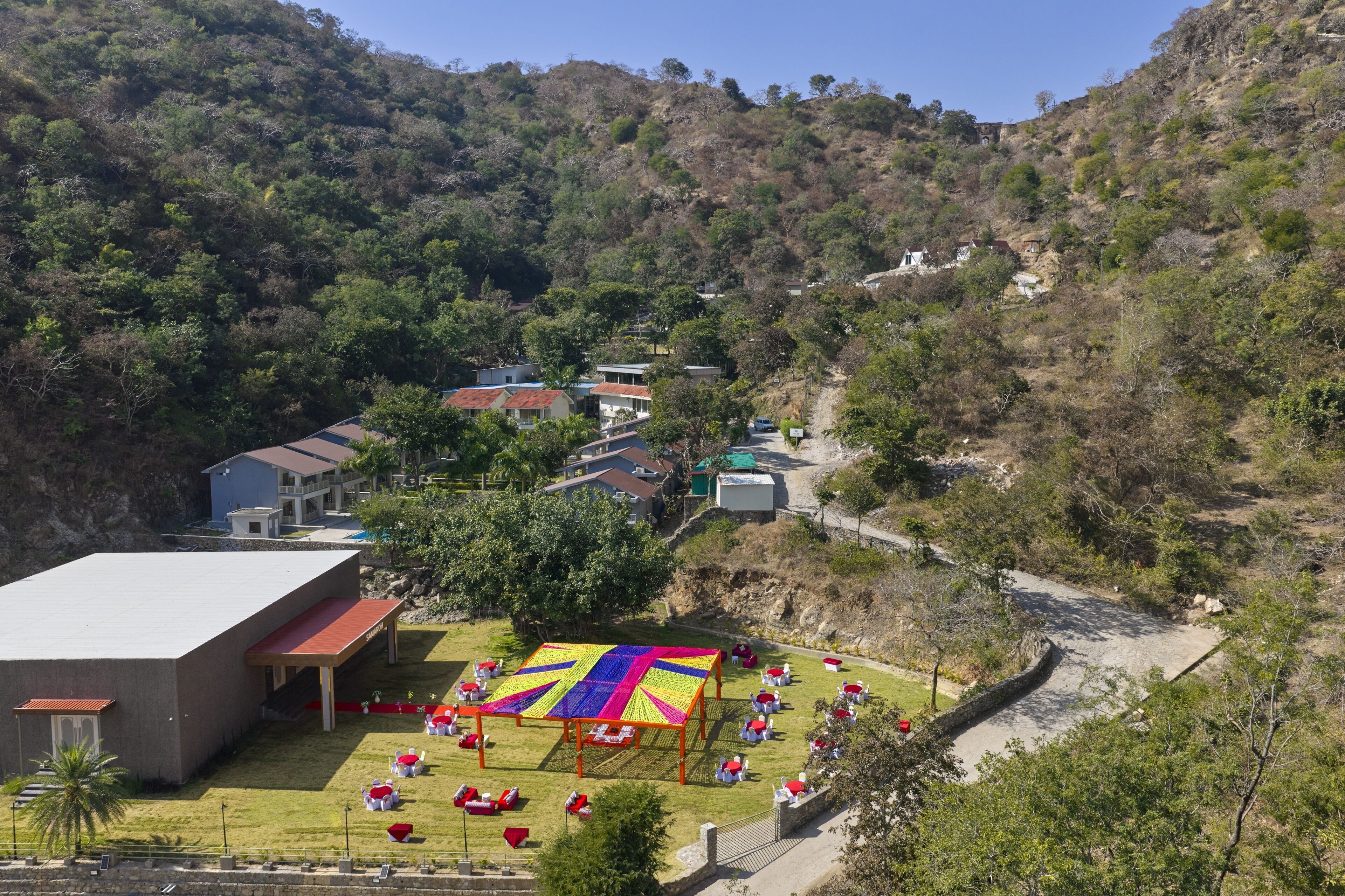 A hillside view of a building with a red roof, a colorful outdoor seating area, and a paved path.