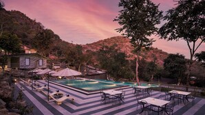 A pool area with lounge chairs and tables, surrounded by trees and hills. - Treat Aranya Resort (Kumbhalgarh)