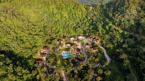 An aerial view of a resort with multiple buildings, a swimming pool, and a winding road through a forested area. - Treat Aranya Resort (Kumbhalgarh)