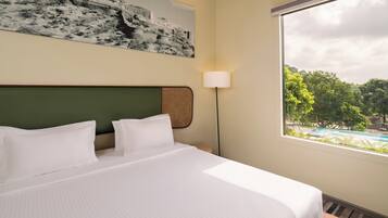 Quarto de hotel com cama, luminária na parede, janela grande com vista para uma piscina e vegetação.
