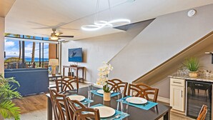 Zona pranzo con tavolo imbandito per quattro persone, ventilatore a soffitto e vista sull'oceano attraverso ampie finestre.