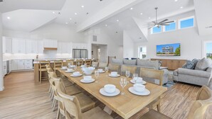 Une cuisine spacieuse avec des meubles blancs, un coin repas avec une table en bois dressée pour six personnes, et un canapé gris dans le salon.