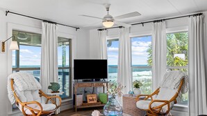 Un salon avec deux fauteuils en osier, une table basse en verre, un téléviseur à écran plat et une vue sur l’océan.