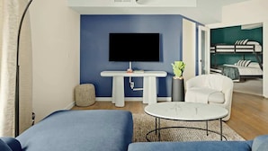 Un salon moderne avec un grand téléviseur à écran plat, une table blanche, une petite table ronde, un fauteuil blanc et, au fond, un lit superposé.
