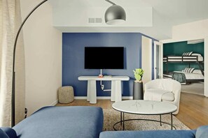 A modern living room with a large flat-screen TV, a white table, a round coffee table, a white armchair, and a bunk bed in the background.