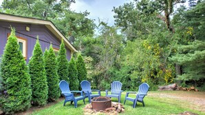Un patio con cuatro sillas Adirondack azules alrededor de un hogar a leña, rodeado por un césped cuidado y altos árboles de hoja perenne.