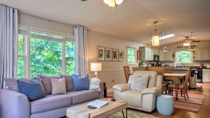 A living room with a sofa, armchair, and coffee table. There is a dining area with a table and chairs. The kitchen is visible in the background.