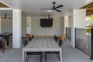 Eine Terrasse im Freien mit einem langen Holztisch und Stühlen, einem Deckenventilator und einem montierten Fernseher.