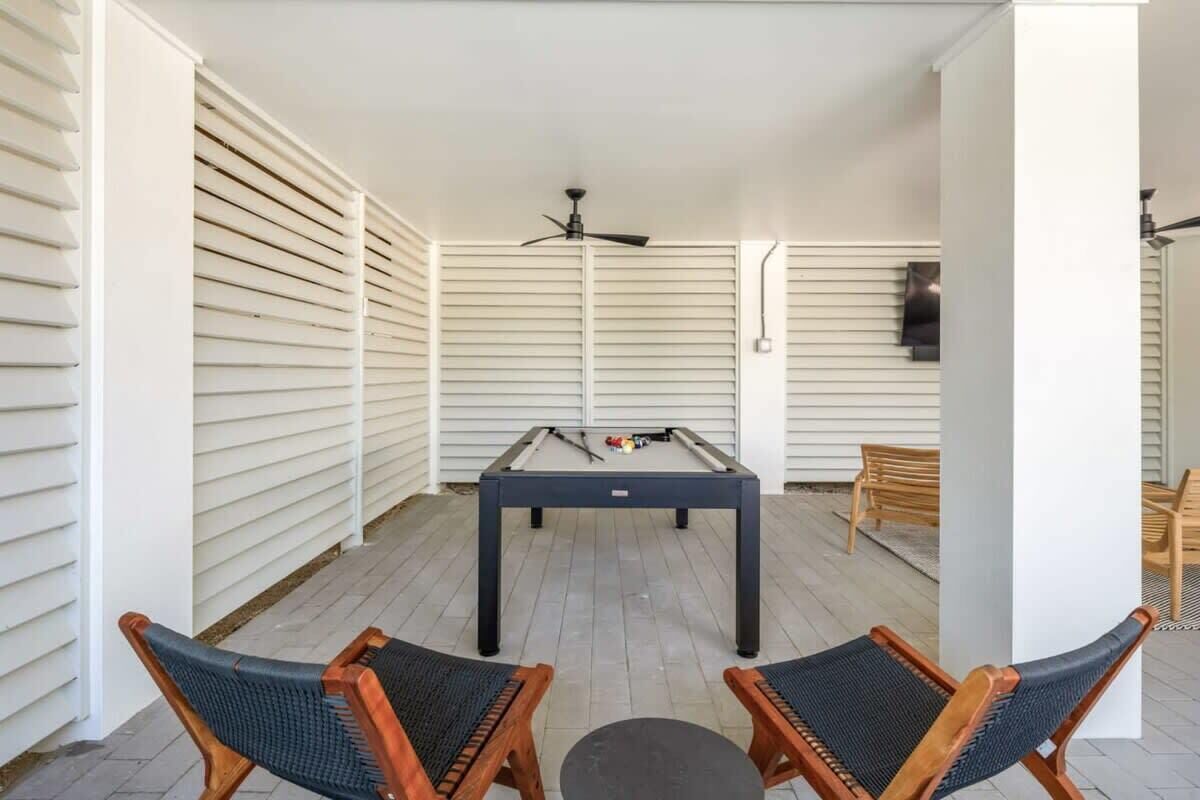 A pool table in a room with white walls and wooden slats, two wicker chairs, and a ceiling fan.