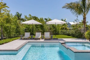 Une piscine avec deux chaises longues et des parasols, entourée de verdure et d’une clôture.