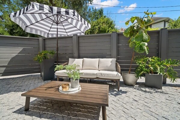 Un espace patio aménagé avec une table en bois, un parasol à rayures, un canapé beige et des plantes en pot.