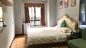 A hotel room with a large bed, a window with curtains, a radiator, and a small table with a green chair.