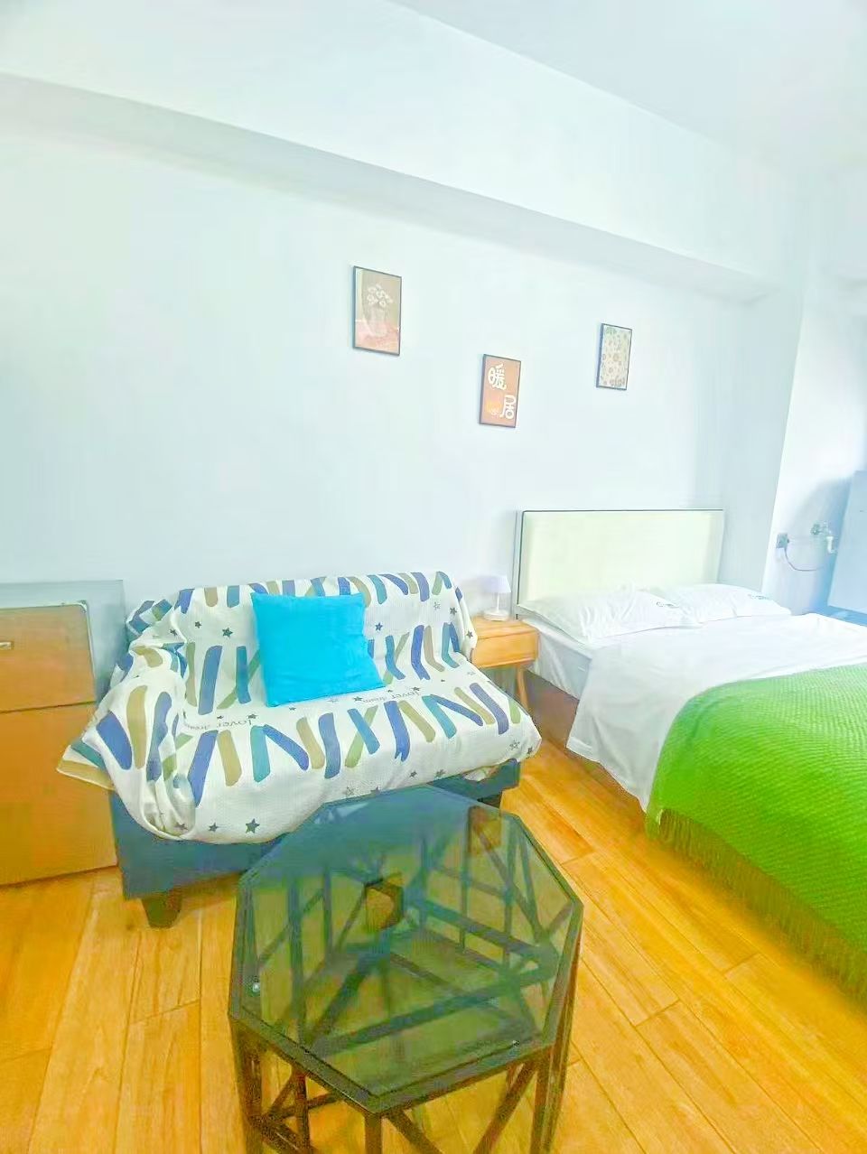 Une petite pièce bien éclairée, avec un canapé, une table en verre, un lit recouvert d’une couverture verte et deux tableaux encadrés accrochés au mur.