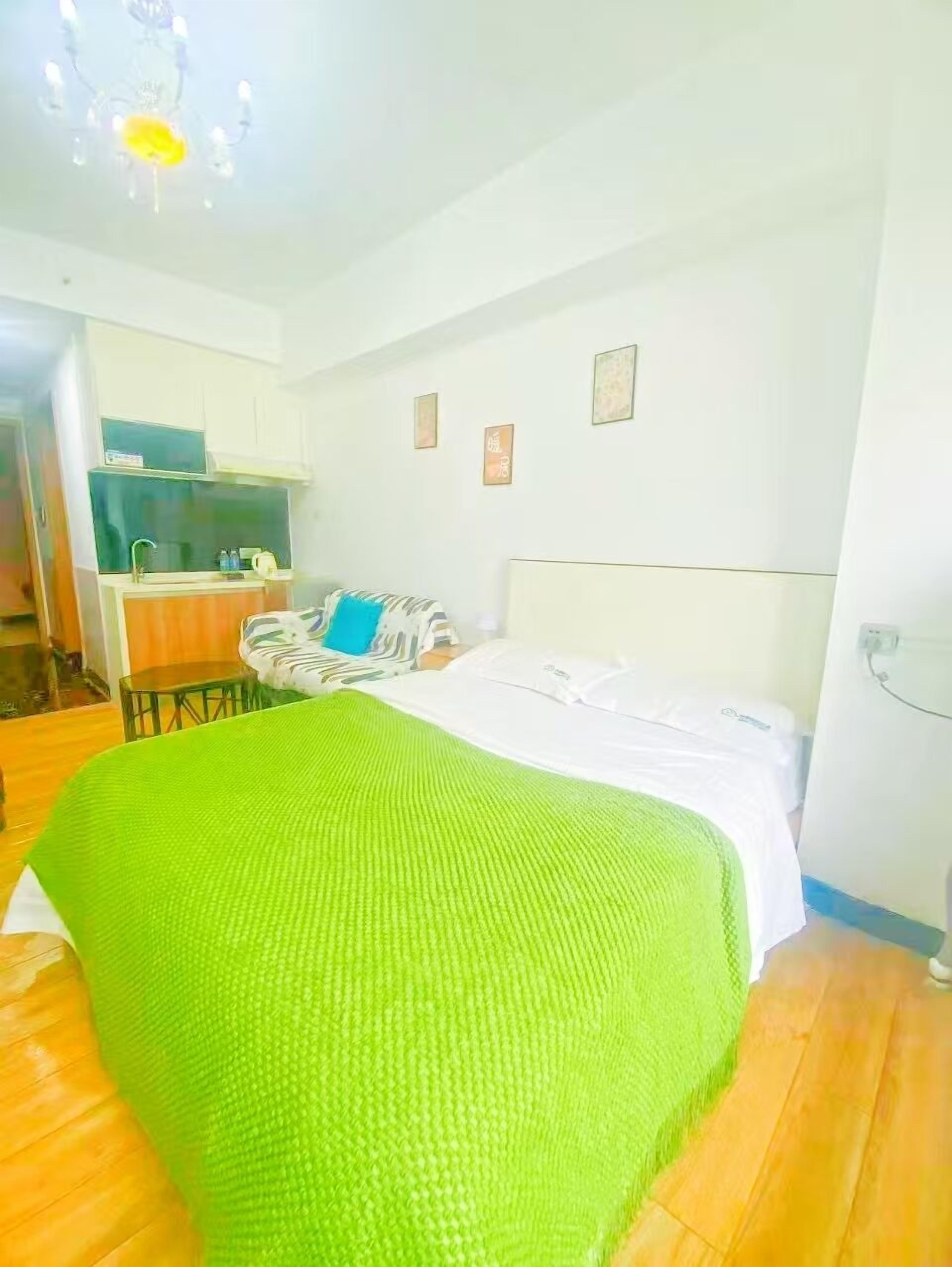 Egy hálószoba, amelyben egy ágy, egy zöld takaró, egy fehér ágyvég és a háttérben egy kis konyhasarok látható.