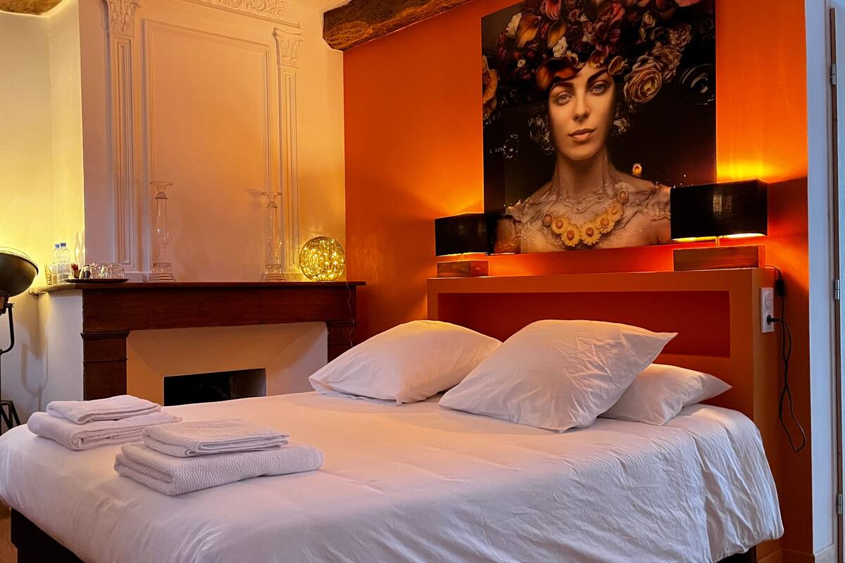 Habitación de hotel con una cama grande, ropa de cama blanca, chimenea, un cuadro en la pared y un escritorio con lámpara.