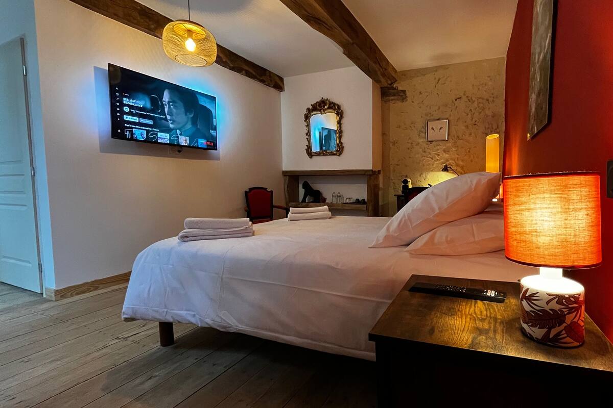 Una cama bien hecha con ropa blanca, un mando a distancia sobre una mesita de noche y un televisor de pared mostrando la interfaz de un servicio de streaming.