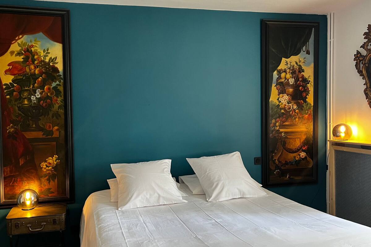 Una habitación de hotel con una cama, dos grandes cuadros florales en las paredes, una mesita de noche con una lámpara y un espejo decorativo.