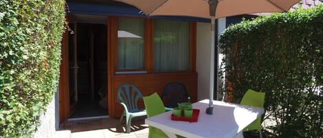 An outdoor patio with a table and chairs, a large umbrella, and a wooden door leading to a room.