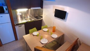 Cocina compacta con fregadero, estufa y un refrigerador pequeño. Hay una mesa de comedor con sillas y un televisor montado en la pared.