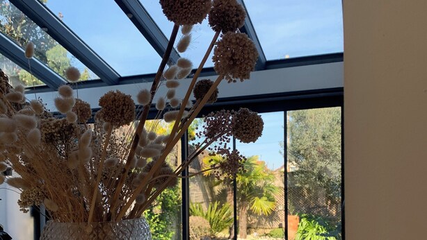 Un jarrón con flores secas sobre una mesa cerca de una ventana que da a un jardín.