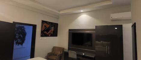 A hotel room with a bed, a desk, a chair, a TV, and an air conditioning unit.