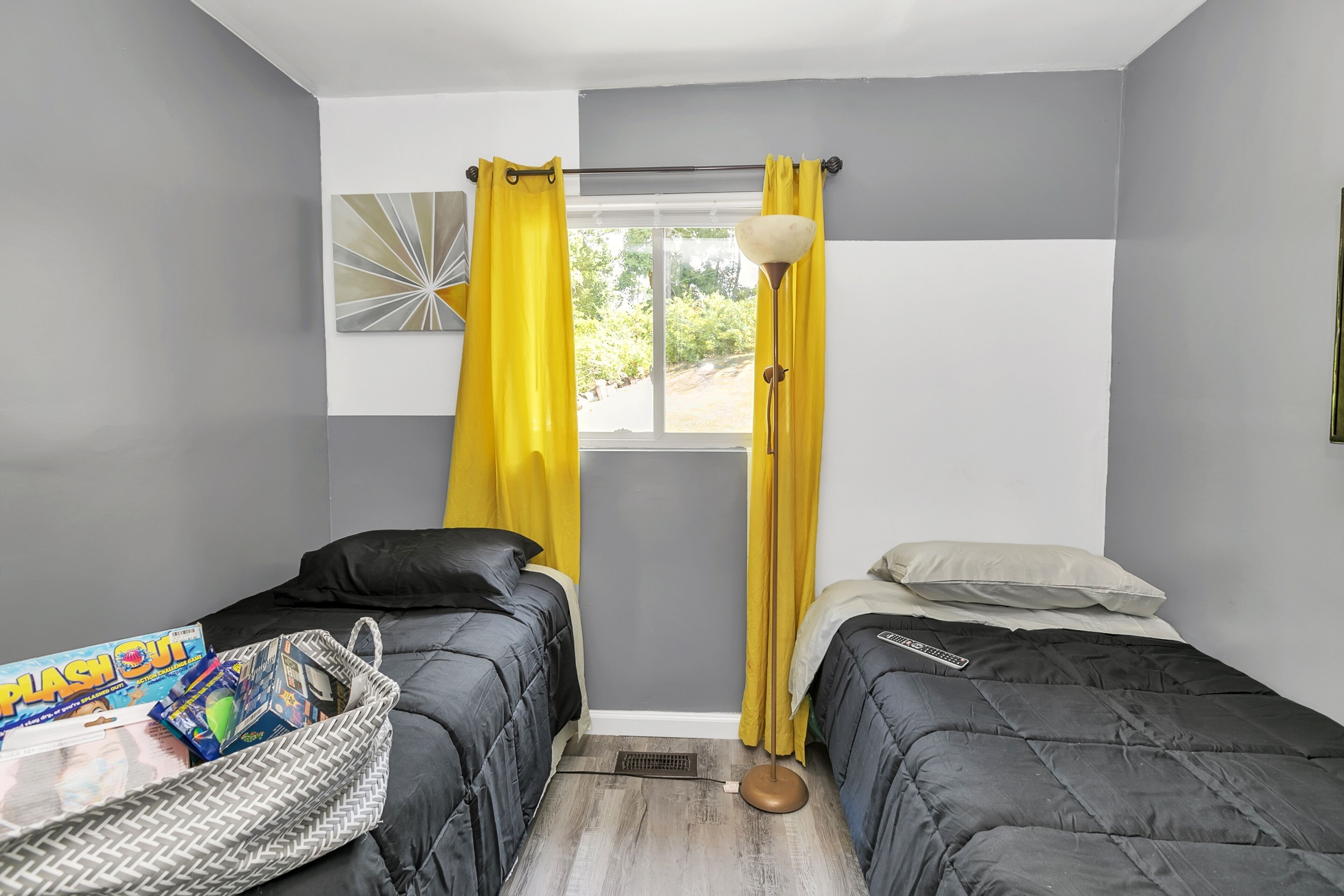 A bedroom with two beds, a window with yellow curtains, a floor lamp, and a basket with magazines.