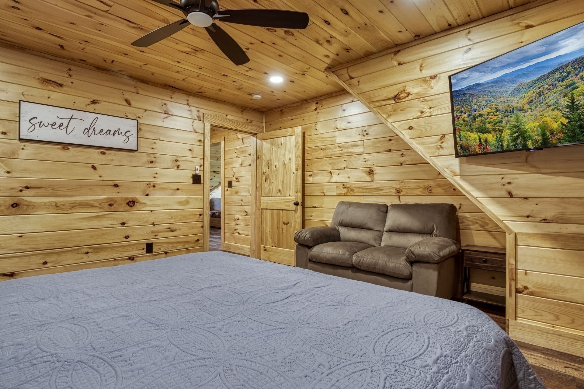 A wooden cabin room with a bed, a leather chair, a ceiling fan, and a wall-mounted TV displaying a scenic view.