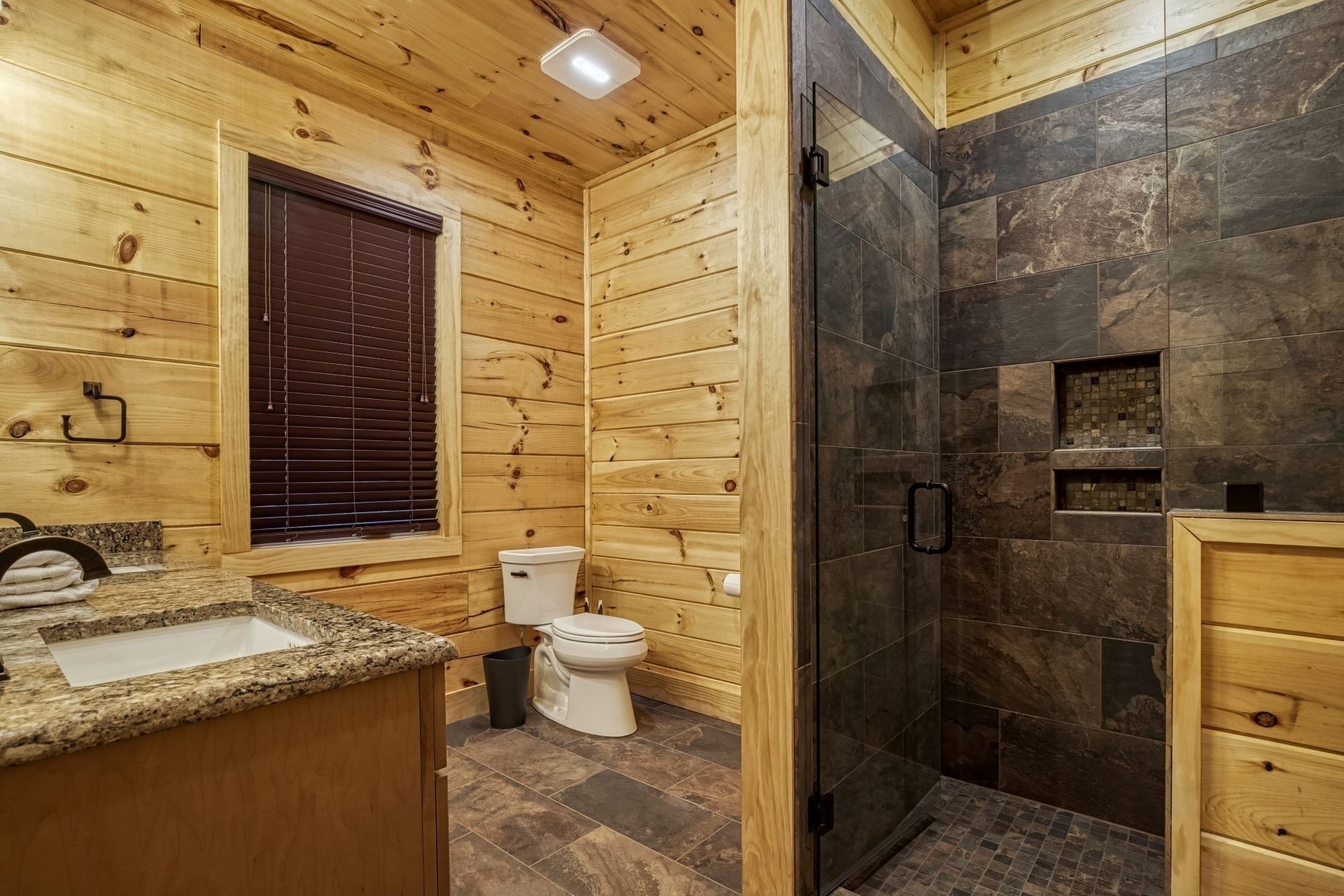 A wooden cabin bathroom with a granite countertop, a toilet, a shower with dark tiles, and a wooden vanity.