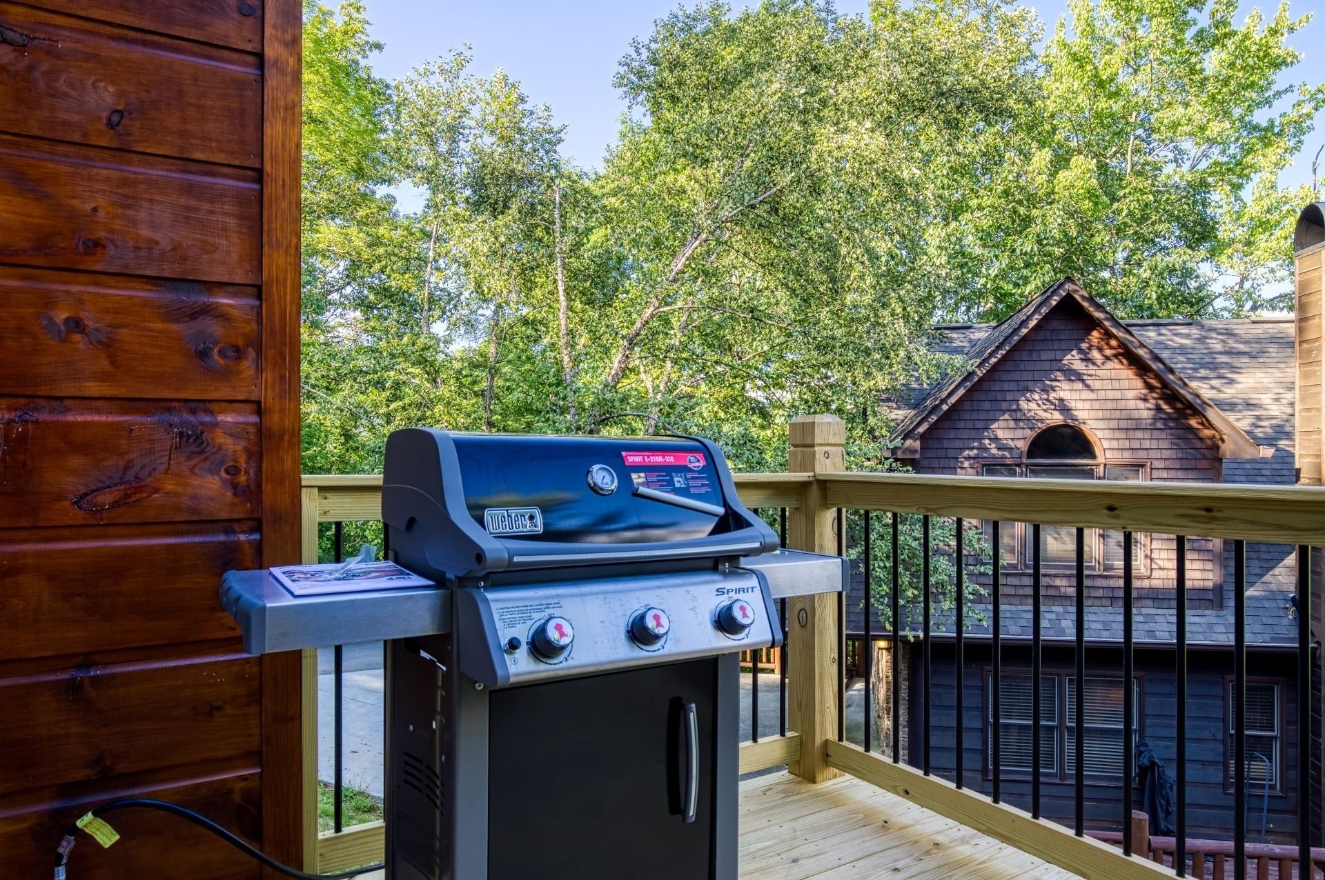 A Weber Spirit gas grill on an outdoor deck with a wooden railing, overlooking a backyard with a wooden building and trees.