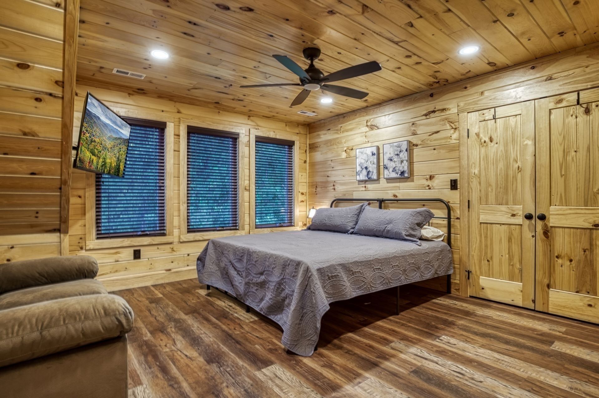Une chambre dans une cabane en bois, avec un lit, un ventilateur de plafond et une grande fenêtre équipée de stores.