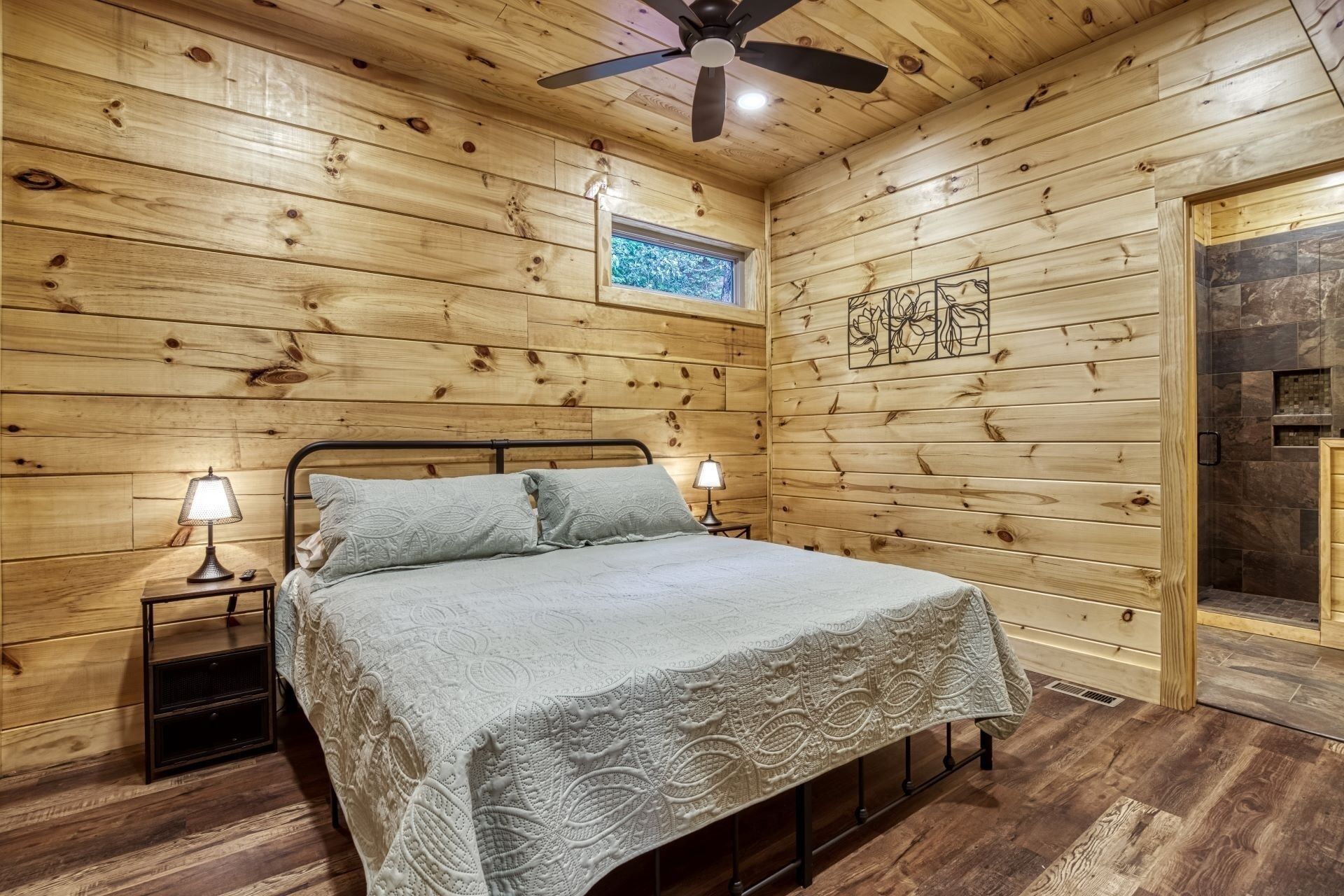 A wooden cabin bedroom with a bed, two bedside lamps, a ceiling fan, and a small window.