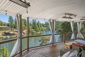 A covered deck with a view of a lake and trees, wooden furniture, and a railing. - Water's Edge - Gorgeous Water Views! by Yosemite Region Resorts (Groveland)