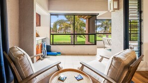 A sunlit patio with two lounge chairs, a small table, and a large window offering a view of a golf course and palm trees. - Big Island Waikoloa Fairways B-214 2 Bedroom Condo (Waikoloa)
