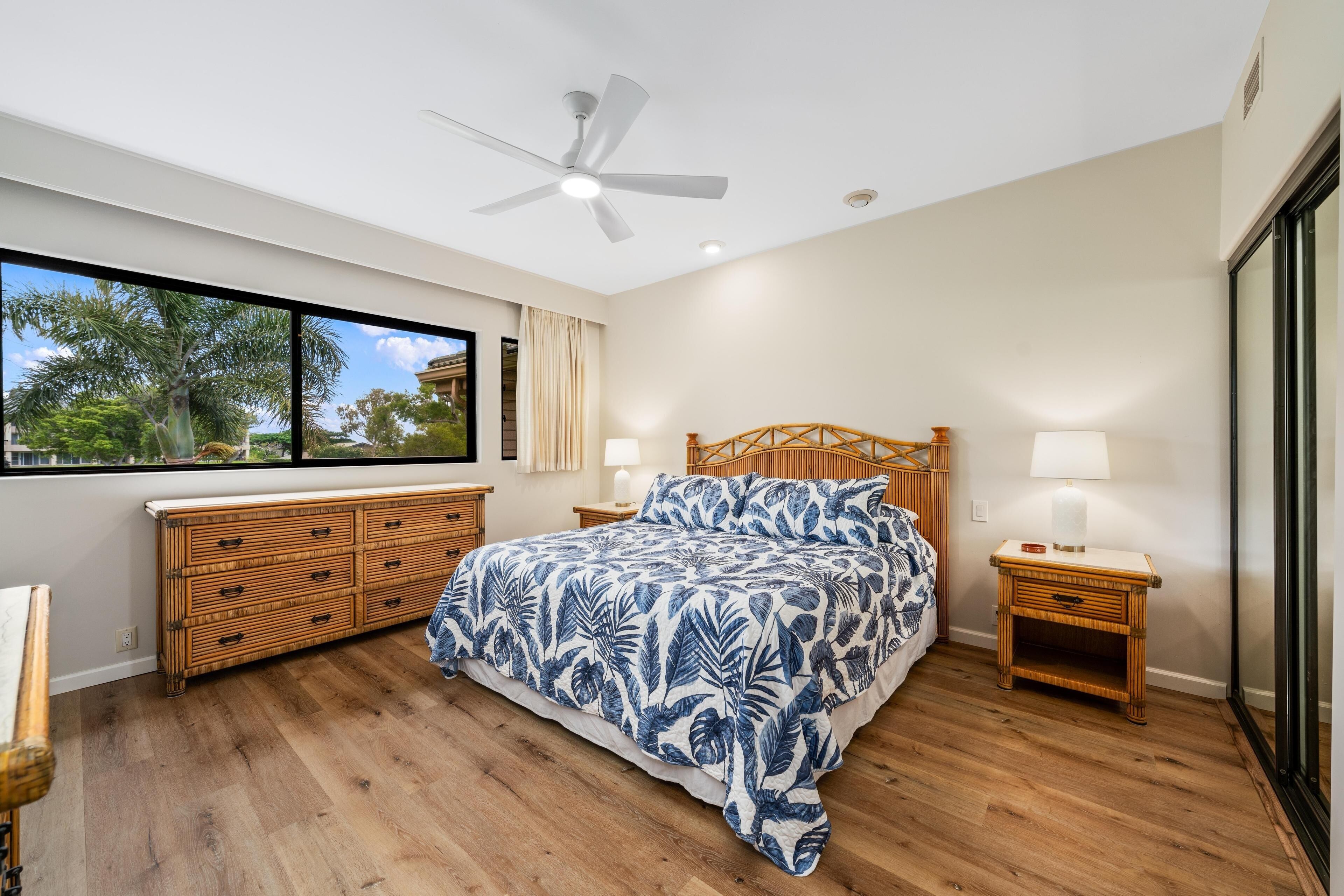A bedroom with a large bed, wooden headboard, a ceiling fan, a window with a view of palm trees, and a wooden nightstand with a lamp.