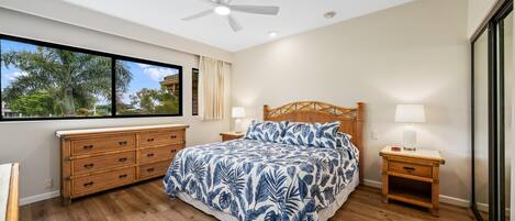 A bedroom with a large bed, wooden headboard, a ceiling fan, a window with a view of palm trees, and a wooden nightstand with a lamp.