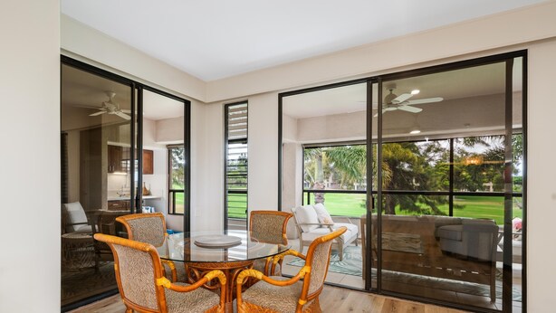 A modern interior with a glass dining table surrounded by wicker chairs, a view of a patio with outdoor furniture, and a ceiling fan. - Big Island Waikoloa Fairways B-214 2 Bedroom Condo (Waikoloa)