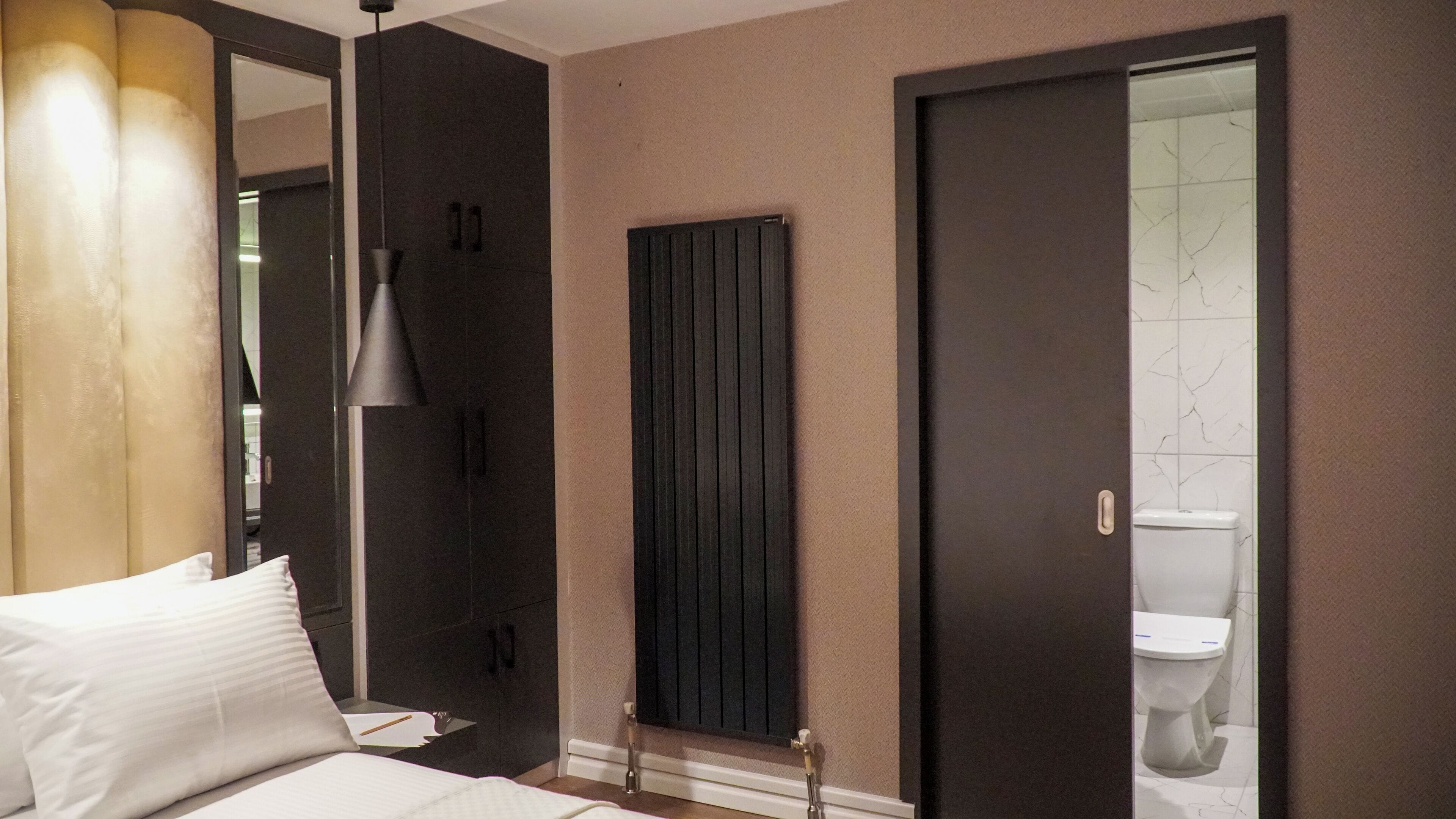 Un dormitorio con cama, una lámpara de noche, una puerta oscura y un baño con inodoro y espejo.