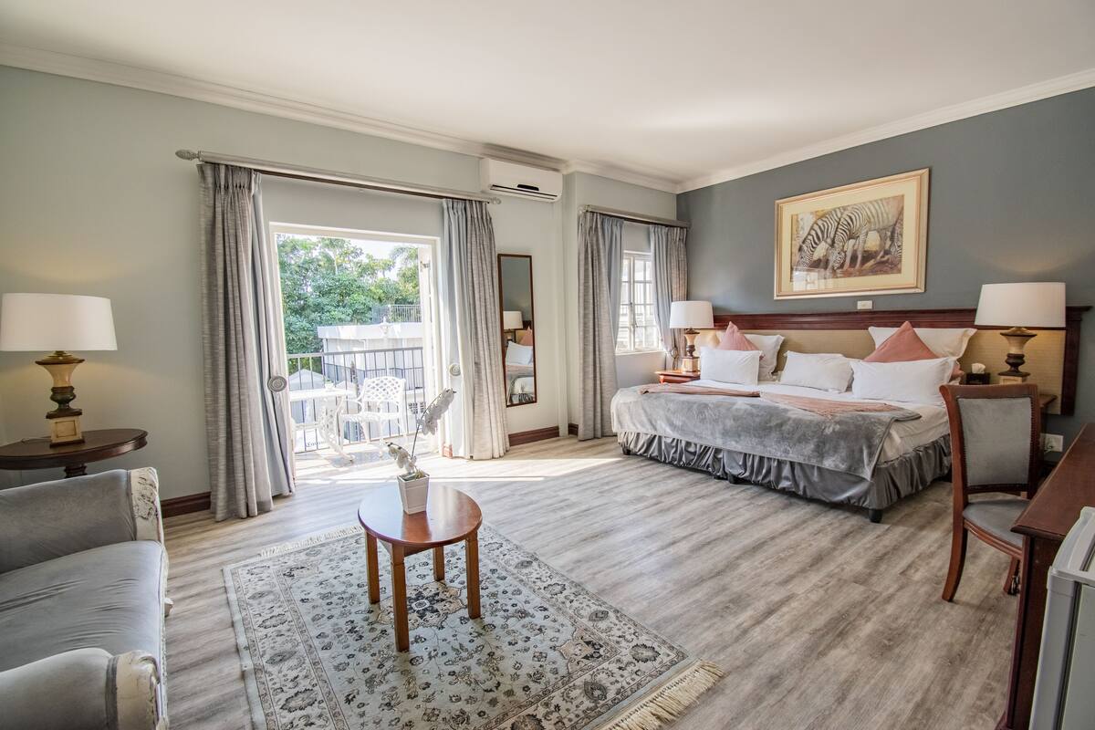 Un dormitorio de hotel con una cama grande, dos mesitas de noche con lámparas, una silla, una pequeña mesa con un florero y vistas al exterior.