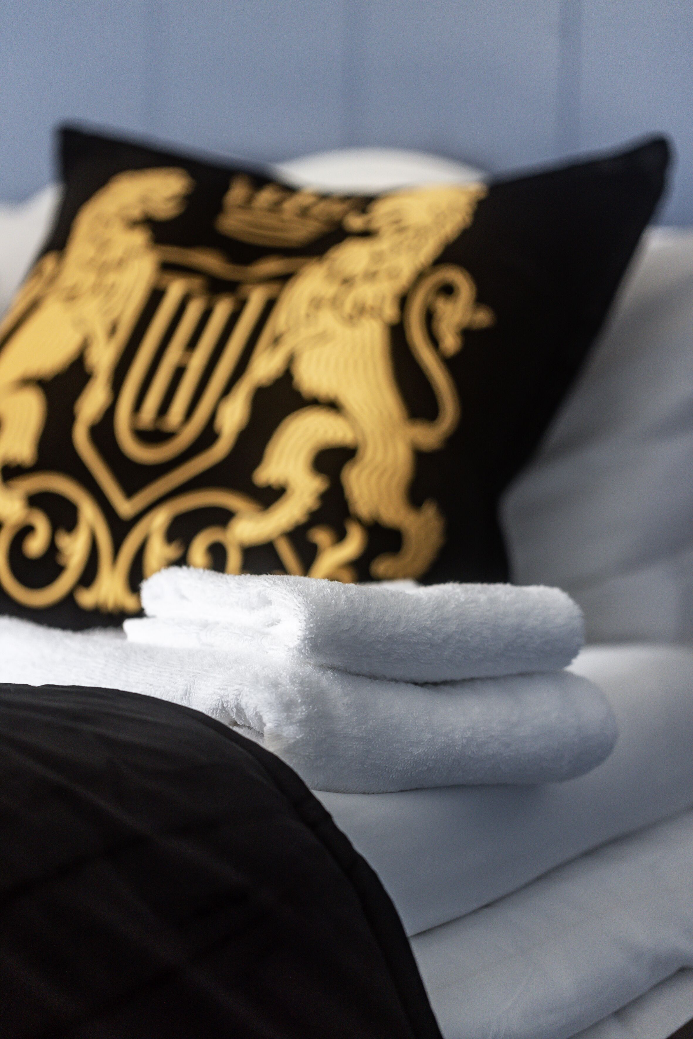 Una toalla blanca doblada con cuidado sobre una cama, con un almohadón negro que lleva un emblema dorado.