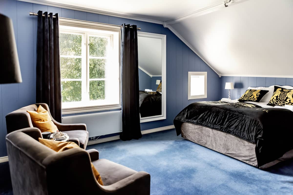 A bedroom with a large bed, a chair, a window with curtains, and a mirror.
