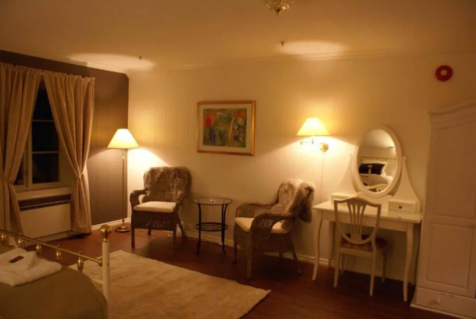 Habitación de hotel con una cama, dos sillas, un escritorio con espejo, una lámpara y un cuadro en la pared.
