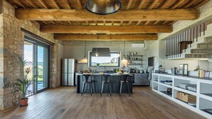 A modern kitchen with a central island, wooden beams, and a large window overlooking a scenic landscape. - Casale The Place 6+2, Emma Villas (San Damiano)