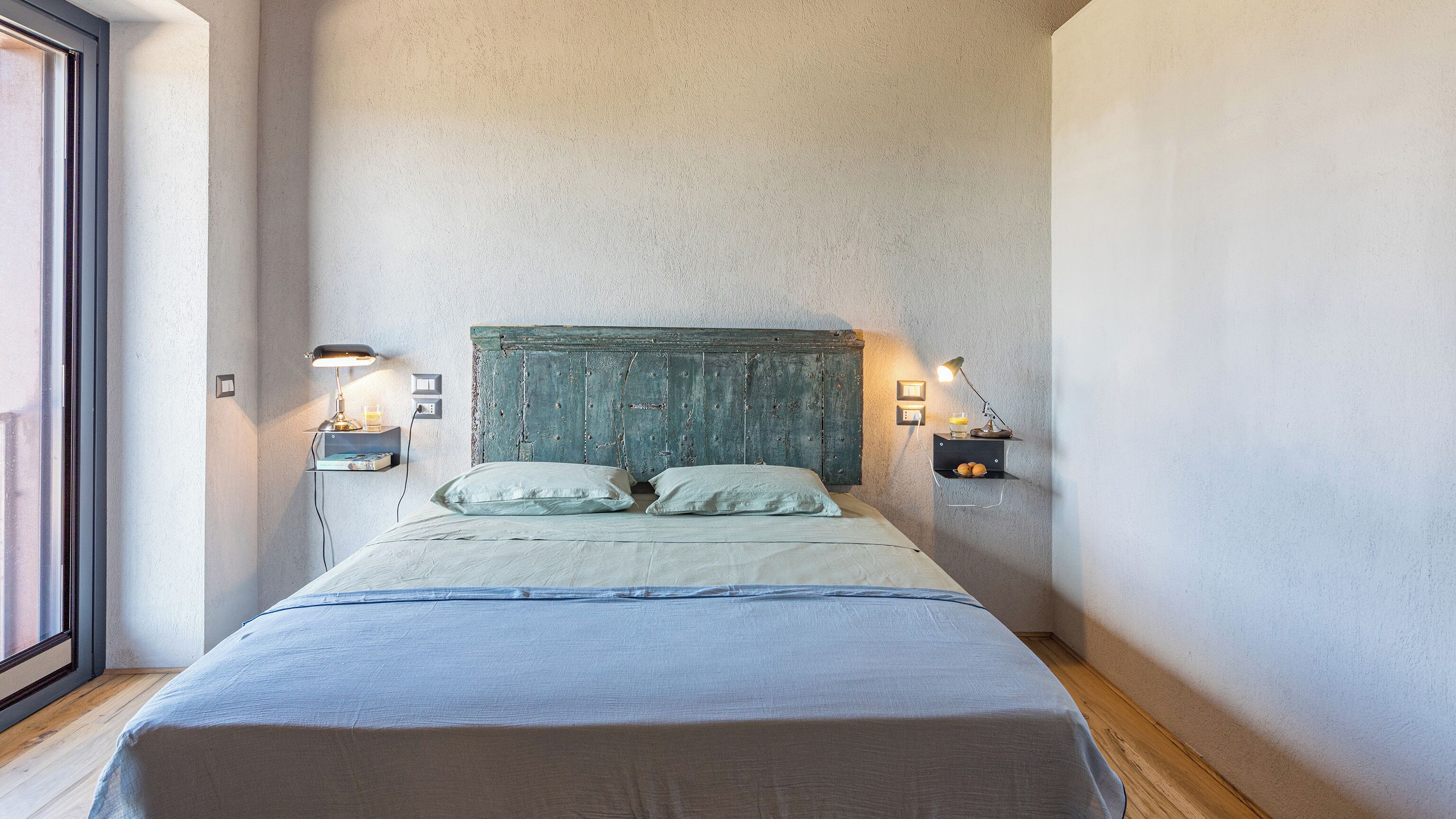 A bedroom with a bed, two bedside tables with lamps, a wall-mounted shelf, and a window.